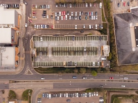 Aerial view of a multi-story parking garage surrounded by parking lots with numerous parked cars. Adjacent to the garage, a busy road runs horizontally across the bottom of the image with several moving vehicles. Surrounding the parking infrastructure are various buildings and green spaces.