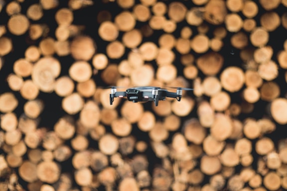 A modern drone hovers in front of a blurred background of stacked cut logs showing the circular patterns of the wood grains.