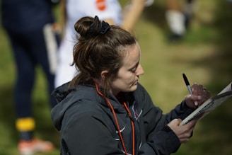 A woman with a bun hairstyle is writing on a clipboard using a black pen. She is wearing a dark jacket and has a red lanyard around her neck. The background is slightly blurred, showing grass and a person in sports clothing.