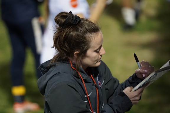 A woman with a bun hairstyle is writing on a clipboard using a black pen. She is wearing a dark jacket and has a red lanyard around her neck. The background is slightly blurred, showing grass and a person in sports clothing.