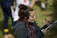 A woman with a bun hairstyle is writing on a clipboard using a black pen. She is wearing a dark jacket and has a red lanyard around her neck. The background is slightly blurred, showing grass and a person in sports clothing.