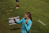 A soccer player on a grassy field appears to be in motion, possibly clapping or gesturing. She is wearing a light blue jersey with an emblem on the sleeve. In the background, a person stands with red socks, and a sign that reads 'Restricted Access' is visible.