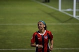 A female soccer player is on a field wearing a red and blue striped jersey, with a logo and number, indicating a team uniform. Her hair is held back with a blue headband, and she appears to be jogging or running with a focused expression. A soccer goal is visible in the background on a green grass field.