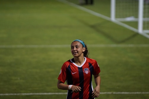 A female soccer player is on a field wearing a red and blue striped jersey, with a logo and number, indicating a team uniform. Her hair is held back with a blue headband, and she appears to be jogging or running with a focused expression. A soccer goal is visible in the background on a green grass field.