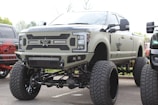 A large, heavily modified pickup truck with an elevated suspension and oversized tires. The truck features custom bumpers, a distinctive grille with a star emblem, and multiple light bars. The body is painted in a matte military green color.