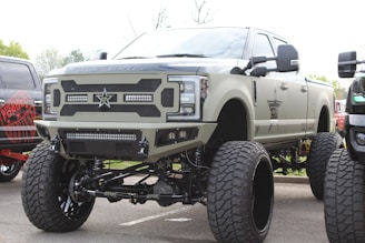 A large, heavily modified pickup truck with an elevated suspension and oversized tires. The truck features custom bumpers, a distinctive grille with a star emblem, and multiple light bars. The body is painted in a matte military green color.