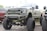 A large, heavily modified pickup truck with an elevated suspension and oversized tires. The truck features custom bumpers, a distinctive grille with a star emblem, and multiple light bars. The body is painted in a matte military green color.
