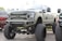 A large, heavily modified pickup truck with an elevated suspension and oversized tires. The truck features custom bumpers, a distinctive grille with a star emblem, and multiple light bars. The body is painted in a matte military green color.