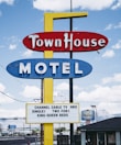 A vintage motel sign with bold red, blue, and yellow colors against a partly cloudy sky. The sign advertises amenities like cable TV and various bed options. In the background, a second smaller sign reads 'Western Inn' with a silhouette of a horse.