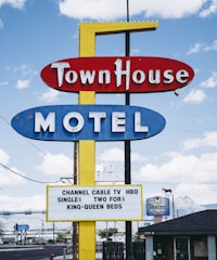 A vintage motel sign with bold red, blue, and yellow colors against a partly cloudy sky. The sign advertises amenities like cable TV and various bed options. In the background, a second smaller sign reads 'Western Inn' with a silhouette of a horse.