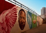 A mural painted on a long wall features a series of vibrant images including large wings, a prominent face, and a hand reaching out. The artwork is set against a backdrop of a cityscape with a clear blue sky, and tall buildings are visible in the distance.