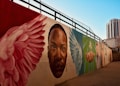 A mural painted on a long wall features a series of vibrant images including large wings, a prominent face, and a hand reaching out. The artwork is set against a backdrop of a cityscape with a clear blue sky, and tall buildings are visible in the distance.