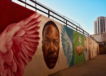 A mural painted on a long wall features a series of vibrant images including large wings, a prominent face, and a hand reaching out. The artwork is set against a backdrop of a cityscape with a clear blue sky, and tall buildings are visible in the distance.