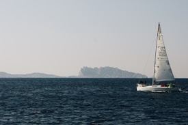A sailboat glides across calm, deep blue waters with two islands visible in the distance under a clear, pale sky. The boat has a white hull and sails, with a few people on board, and the sail displays various numbers and letters.