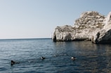 A group of divers swims in a calm, deep blue sea near a rocky cliff. In the background, a sailboat can be seen on the horizon, set against a clear sky.