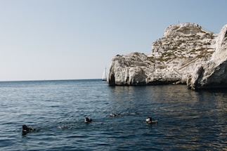 A group of divers swims in a calm, deep blue sea near a rocky cliff. In the background, a sailboat can be seen on the horizon, set against a clear sky.