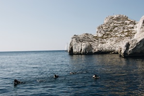 A group of divers swims in a calm, deep blue sea near a rocky cliff. In the background, a sailboat can be seen on the horizon, set against a clear sky.