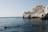 A group of divers swims in a calm, deep blue sea near a rocky cliff. In the background, a sailboat can be seen on the horizon, set against a clear sky.