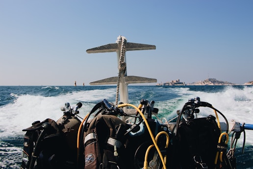 A view from a boat with multiple scuba diving tanks and gear visible at the back. The ocean is choppy with waves, and there is a small island or coastal area in the distance. The sky is clear and sunny.