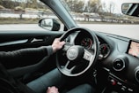 A person is driving a car, with their left hand on the steering wheel. The dashboard includes a speedometer and a small display showing a GPS map. The road outside is visible through the windshield, along with blurred trees and a guardrail, indicating the vehicle is in motion. A side mirror reflects part of the surrounding scene.