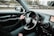 A person is driving a car, with their left hand on the steering wheel. The dashboard includes a speedometer and a small display showing a GPS map. The road outside is visible through the windshield, along with blurred trees and a guardrail, indicating the vehicle is in motion. A side mirror reflects part of the surrounding scene.