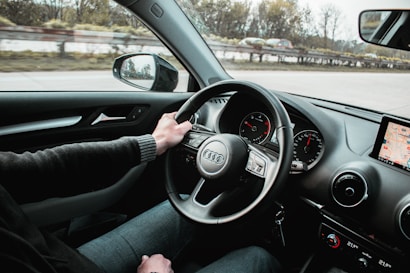 A person is driving a car, with their left hand on the steering wheel. The dashboard includes a speedometer and a small display showing a GPS map. The road outside is visible through the windshield, along with blurred trees and a guardrail, indicating the vehicle is in motion. A side mirror reflects part of the surrounding scene.