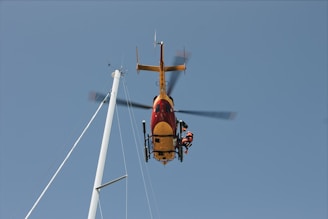A helicopter is hovering in the air with a person on a rescue line below it. The helicopter is red and yellow and is positioned near a tall mast against a clear blue sky.