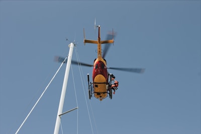A helicopter is hovering in the air with a person on a rescue line below it. The helicopter is red and yellow and is positioned near a tall mast against a clear blue sky.