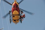 A helicopter captured from below, with a rescue worker in a harness attached to it. The helicopter is brightly colored in red and yellow. The worker is wearing a red and black uniform with a helmet, indicating a rescue operation.