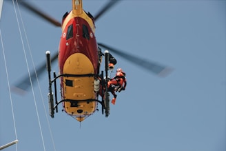 A helicopter captured from below, with a rescue worker in a harness attached to it. The helicopter is brightly colored in red and yellow. The worker is wearing a red and black uniform with a helmet, indicating a rescue operation.