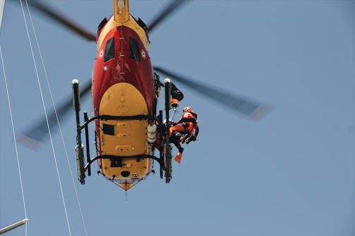 A helicopter captured from below, with a rescue worker in a harness attached to it. The helicopter is brightly colored in red and yellow. The worker is wearing a red and black uniform with a helmet, indicating a rescue operation.