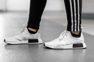 A person is wearing white Adidas sneakers with black accents, paired with black pants featuring white stripes running down the sides. The photo captures them from the mid-shin down, standing on a smooth, gray floor in what appears to be an indoor setting.