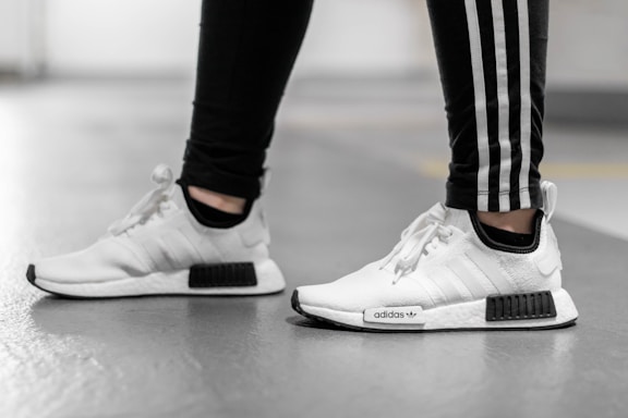A person is wearing white Adidas sneakers with black accents, paired with black pants featuring white stripes running down the sides. The photo captures them from the mid-shin down, standing on a smooth, gray floor in what appears to be an indoor setting.