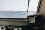 A nautical control panel featuring a compass and a navigation screen is seen through a ship's window with a view of the open sea and horizon under a clear sky.