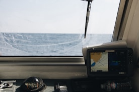 A nautical control panel featuring a compass and a navigation screen is seen through a ship's window with a view of the open sea and horizon under a clear sky.