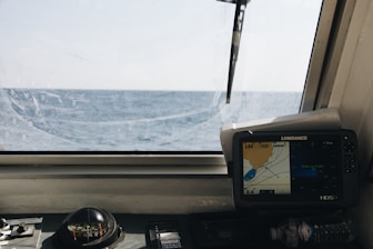 A nautical control panel featuring a compass and a navigation screen is seen through a ship's window with a view of the open sea and horizon under a clear sky.