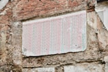 A weathered brick wall features a large section covered with a faded, patterned wallpaper. The wallpaper appears old and tattered, with its colors dulled over time. The surrounding bricks are irregular and show signs of age and erosion.