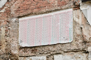 A weathered brick wall features a large section covered with a faded, patterned wallpaper. The wallpaper appears old and tattered, with its colors dulled over time. The surrounding bricks are irregular and show signs of age and erosion.