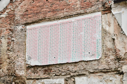 A weathered brick wall features a large section covered with a faded, patterned wallpaper. The wallpaper appears old and tattered, with its colors dulled over time. The surrounding bricks are irregular and show signs of age and erosion.