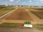 An expansive farm scene features a large field of crops with a green combine harvester working alongside a red truck with a large white trailer. The landscape extends across flat, fertile farmland, with multiple sections of fields in various shades of brown and green. Buildings and silos are visible in the distant background under a sky with scattered clouds.