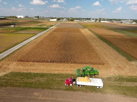 An expansive farm scene features a large field of crops with a green combine harvester working alongside a red truck with a large white trailer. The landscape extends across flat, fertile farmland, with multiple sections of fields in various shades of brown and green. Buildings and silos are visible in the distant background under a sky with scattered clouds.