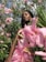 A person with braided hair stands amidst lush greenery and vibrant pink flowers. The sunlight highlights their confident expression, creating a striking contrast between the softness of the flowers and their bold presence.
