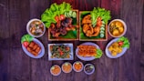 A variety of dishes are arranged on a dark wooden table, featuring grilled and fried chicken pieces garnished with fresh lettuce, tomatoes, and cucumber slices. There are bowls of soup, fried spring rolls, a plate of sautéed vegetables with sliced chilies, skewered shrimp, tofu, and small bowls of dipping sauces with lime. The presentation is vibrant and appetizing.