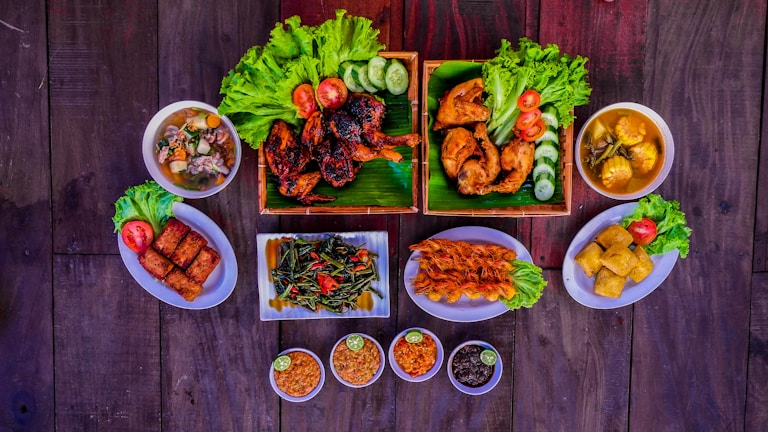 A variety of dishes are arranged on a dark wooden table, featuring grilled and fried chicken pieces garnished with fresh lettuce, tomatoes, and cucumber slices. There are bowls of soup, fried spring rolls, a plate of sautéed vegetables with sliced chilies, skewered shrimp, tofu, and small bowls of dipping sauces with lime. The presentation is vibrant and appetizing.