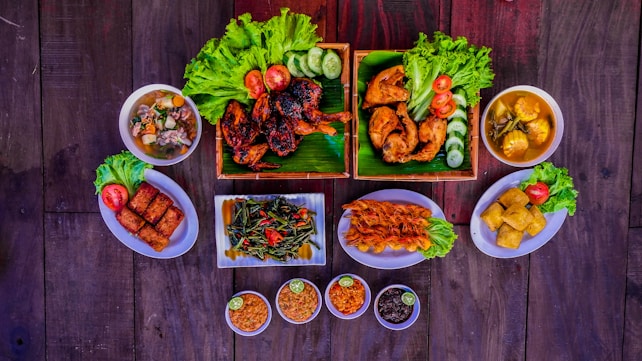 A variety of dishes are arranged on a dark wooden table, featuring grilled and fried chicken pieces garnished with fresh lettuce, tomatoes, and cucumber slices. There are bowls of soup, fried spring rolls, a plate of saut&eacute;ed vegetables with sliced chilies, skewered shrimp, tofu, and small bowls of dipping sauces with lime. The presentation is vibrant and appetizing.