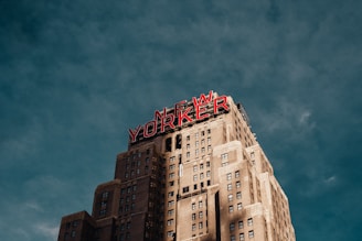 A tall building with a classic architectural style features a prominent red neon sign displaying the words 'New Yorker' against a moody, cloudy sky. The building's facade is detailed with rectangular windows and a pattern of lighter brickwork.
