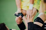 A close-up of a sports interaction where one person in a yellow uniform helps another wearing black leg gear. The person in yellow is gripping the leg of the other individual, providing support. The background appears to be a green sports field.