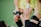 A close-up of a sports interaction where one person in a yellow uniform helps another wearing black leg gear. The person in yellow is gripping the leg of the other individual, providing support. The background appears to be a green sports field.
