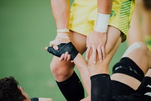 A close-up of a sports interaction where one person in a yellow uniform helps another wearing black leg gear. The person in yellow is gripping the leg of the other individual, providing support. The background appears to be a green sports field.