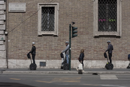 Three people on Segways are lined up next to a brick wall with a street sign labeled 'Via di S. Marco'. A pedestrian with a bag walks past them. They are on the sidewalk next to a traffic light.